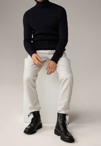 Maglione a collo alto a coste blu navy, jeans slim-fit color crema e stivali neri con lacci e suola spessa, seduto su un piedistallo bianco.