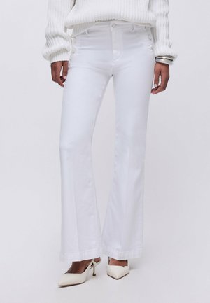 Persona con pantaloni bianchi a zampa, maglione a maglia bianco, scarpe bianche con tacco e braccialetti argentati al polso sinistro, in piedi davanti a uno sfondo semplice.