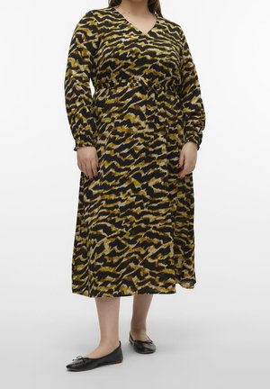 Osoba plus size w długiej sukience z długim rękawem i dekoltem w serek, o długości midi, z nadrukiem tygrysa w kolorach czarnym, brązowym i beżowym, w towarzystwie czarnych balerinek.