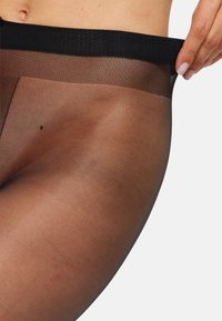 Nærbillede af en person, der trækker sorte, gennemsigtige strømpebukser op med en sort talje, som viser hudfarve og hånddetaljer.