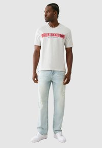 Pilkos medvilninės t-shirt su raudonu "TRUE RELIGION" logotipu, derinama su šviesiai mėlynomis suplyšusiomis džinsomis ir baltomis sportbačiais, stovintis prieš neutralų foną.