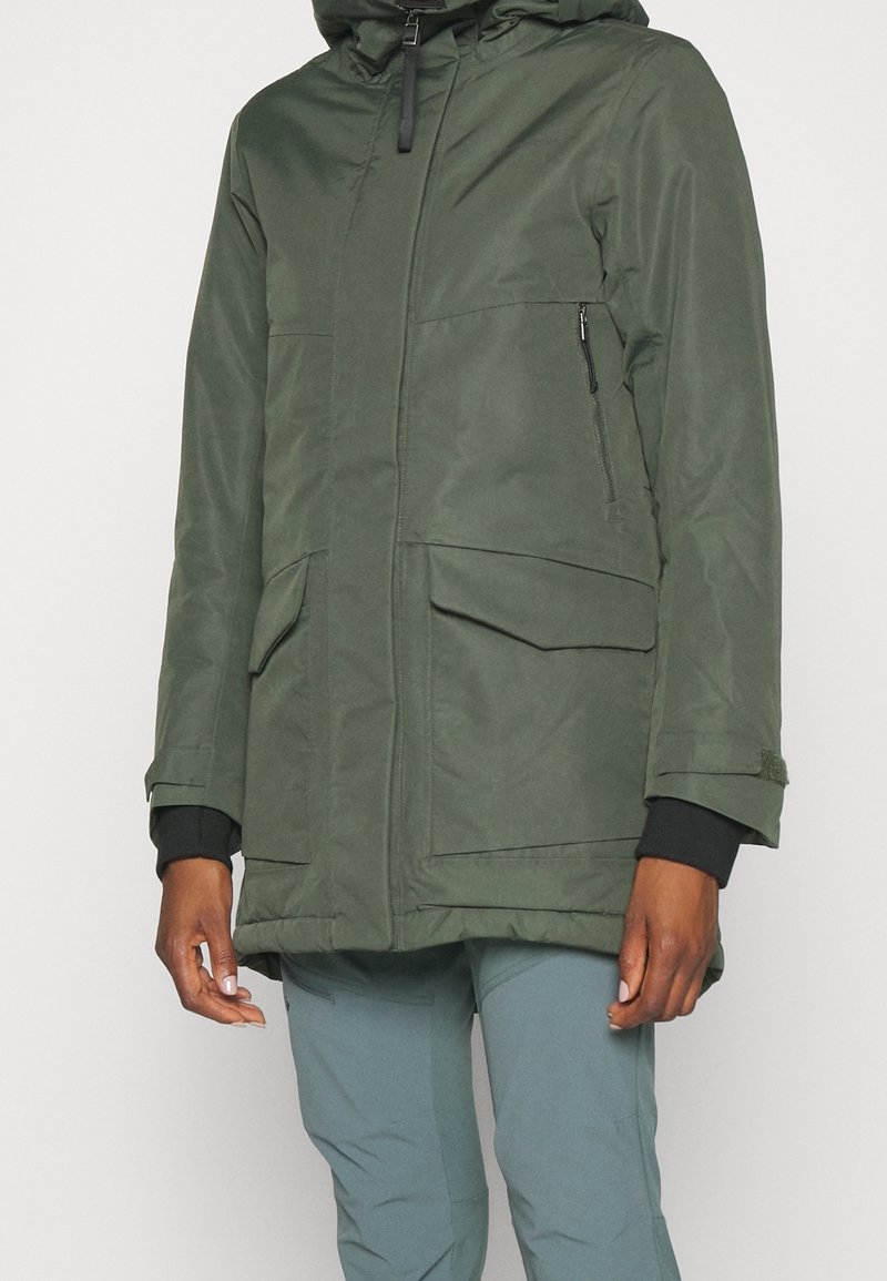 Personne portant une veste d'hiver verte à capuche avec rabat frontal et poches zippées, et un pantalon bleu-gris sur fond blanc uni.