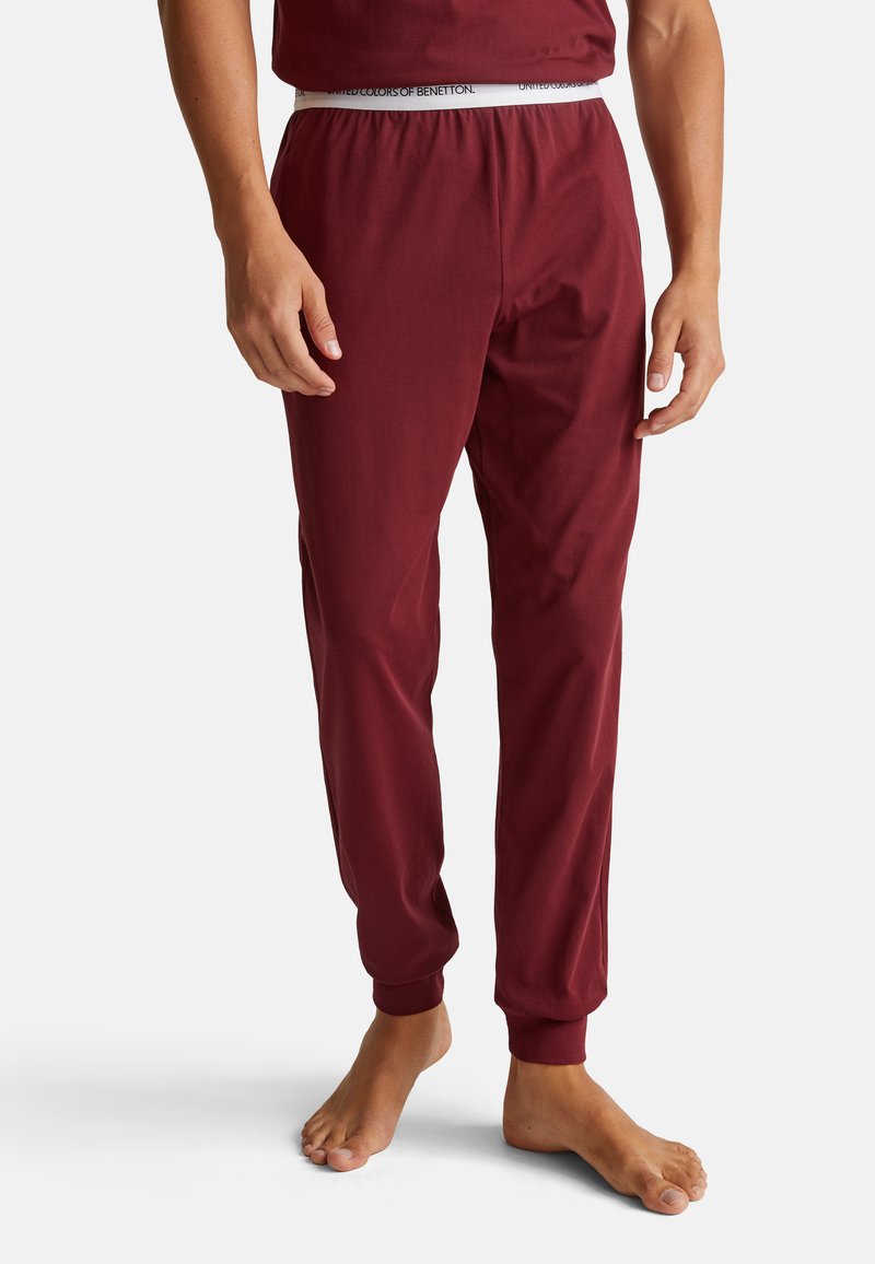 Muž stojící bos ve vínových joggingových kalhotách a odpovídajícím triku na bílém pozadí.