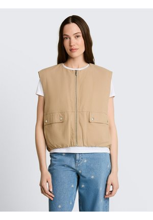Eine junge Frau trägt eine beige ärmellose Weste mit Reißverschluss über einem weißen T-Shirt und blauen Jeans mit kleinen weißen, bestickten Blumenmustern.