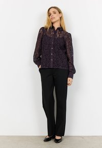 Kvinna klädd i en mörklila spetsblus med långa ärmar, svarta byxor med rak ben, och svarta lackskor, stående mot en vit vägg.