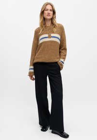 Bruine donzige trui met witte en blauwe horizontale strepen, met een drie-knopen sluiting; gedragen met zwarte streepjumpantalons en zwarte schoenen.