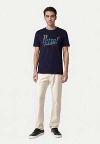 Marineblå bomulds T-shirt med "Petrol Industries" grafik i teal og guld, parret med lyse beige bukser og sorte sneakers.