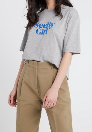 Žena v šedém tričku SeeByGirl zastrčeném do vysokých béžových kalhot, stojící před jednoduchým světlým pozadím.