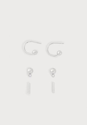 Deux paires de boucles d'oreilles en argent sur un fond clair, une paire avec des anneaux ouverts et des extrémités en boule, l'autre avec des clous ronds et de petites barres pendantes.