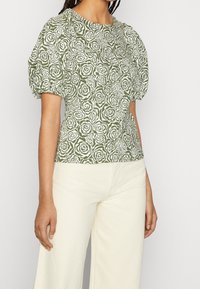 Zielona bluzka w kwiatowy wzór z bufkami, ozdobiona białymi różami. Połączona z jasnobeżowymi, szerokimi spodniami.