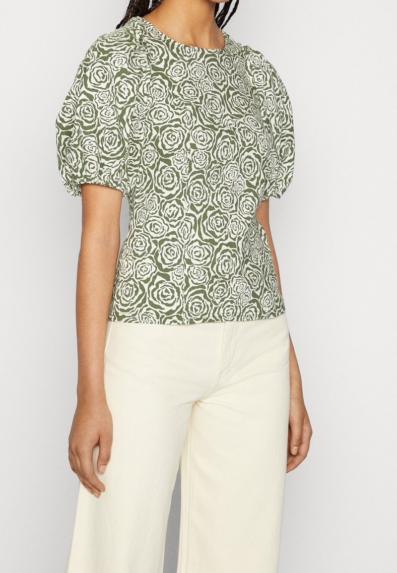 Zielona bluzka w kwiatowy wzór z bufkami, ozdobiona białymi różami. Połączona z jasnobeżowymi, szerokimi spodniami.