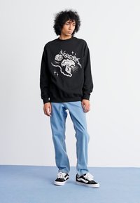 Felpa nera con un design grafico bianco, abbinata a jeans blu chiaro e sneakers bianco e nere. Sfondo semplice.