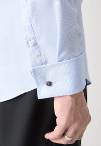 Manžeta světle modré texturované košile s bílým knoflíkem a tmavým detailem. Košile má čtvercový vzor a design s přehnutou manžetou.