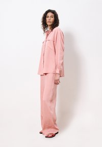 Pink satin two-piece set featuring a long-sleeve button-up shirt with chest pockets and wide-leg pants. Model wears red flip-flops.