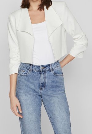 Frau trägt eine kurze weiße Jacke über einem weißen Oberteil, kombiniert mit hoch taillierten hellblauen Jeans, steht mit einer Hand in der Hüfte.