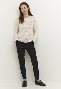 Top in pizzo a maniche lunghe di colore crema, abbinato a pantaloni neri su misura. Mocassini neri completano l'outfit. Visibili il colore solido e i dettagli testurizzati.