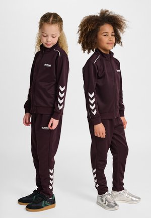Bordeauxfarbene Trainingsanzüge aus weichem Stoff, mit weißen Chevronstreifen an den Ärmeln und den Seiten der Hose. Logo auf der Brust, kombiniert mit schwarzen und silbernen Sneakers.