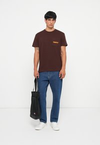 Tricou din bumbac maro cu buzunar pe piept, având un logo portocaliu, asortat cu blugi albaștri și adidași albi. Modelul ține o geantă de tote neagră.