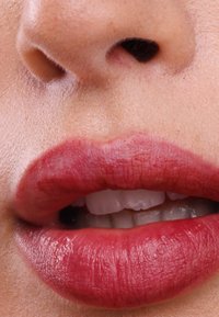 Primer plano de labios con un acabado rojo brillante, mostrando textura y un sutil degradado. Se ven los dientes entre labios ligeramente entreabiertos.