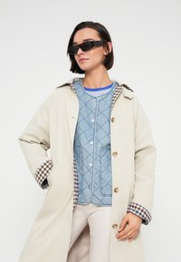 Μπεζ αδιάβροχο παλτό με επένδυση καρό, φορεμένο πάνω από ένα στρώμα γαλάζιου τζιν με καπιτονέ υφή. Μαύρα γυαλιά ηλίου συμπληρώνουν την εμφάνιση.