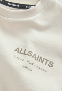 Krémové tričko z látky s kulatým výstřihem. Na přední straně je vytištěn text "ALLSAINTS" a "LONDON" v šedé barvě.