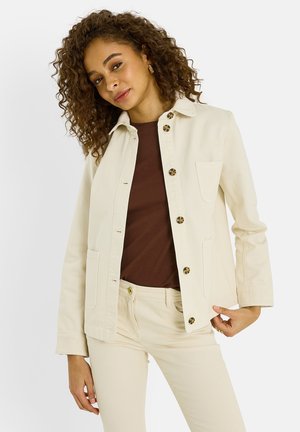 Mujer con cabello rizado, vestida con chaqueta beige abotonada, pantalones a juego y top marrón oscuro, sobre un fondo claro y liso.