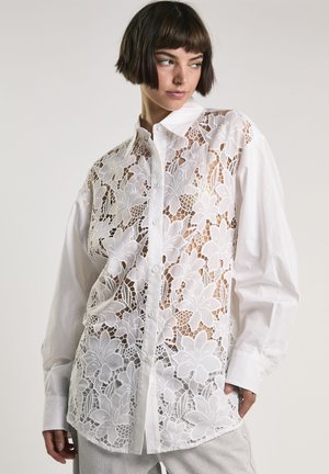 Donna che indossa una camicia bianca con bottoni, sovrapposizione in pizzo floreale sul davanti e maniche lunghe semplici, guarda di lato.