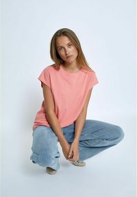 Rožinė medvilninė marškinė su trumpomis rankovėmis, apvaliu iškirpimu, deranti su laisvais mėlynais džinsais. Modelis keliuose, dėvintis tekstūruotus plokščius batus.