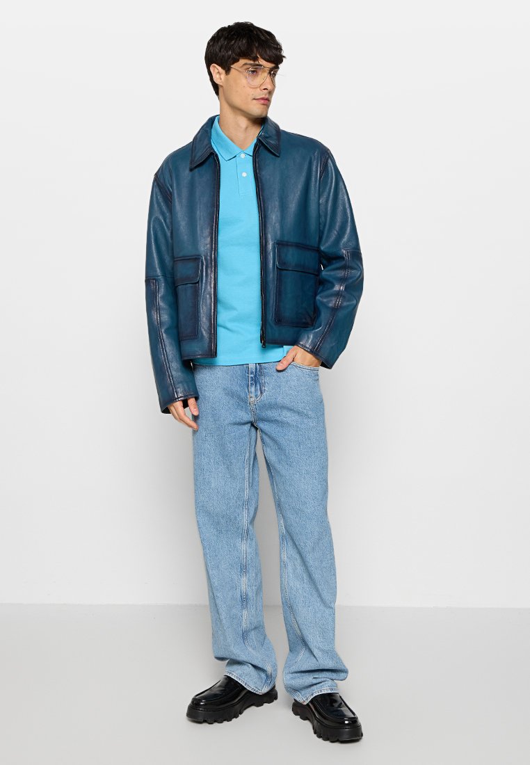 Blauwe leren jas met twee voorzakken over een lichtblauw polo shirt. Lichtblauwe jeans en zwarte platform schoenen maken de outfit compleet.