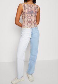 Hnědý a béžový tie-dye bezrukávový top spárovaný se světle modrými a bílými rozparkovými džíny, doplněný béžovými sportovními botami. Detail řetízku na krku.