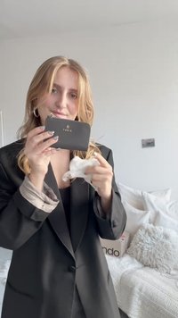 Carteira de couro preta com logótipo dourado, formato retangular, textura lisa, segurada em frente a uma pessoa vestindo um blazer preto.