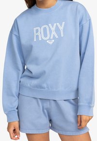 Felpa azzurra con logo "ROXY" bianco, caratterizzata da un colletto tondo, polsini a costine e una vestibilità ampia, abbinata a pantaloni corti coordinati.