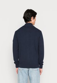 Marineblauwe pullover trui met een geribbelde kraag en manchetten, met een textuur gebreid ontwerp. Weergegeven van de achterkant met lichtblauwe spijkerbroek.