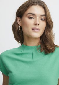 Top verde texturat cu guler înalt, având detalii plisate. Modelul are păr ondulat maro și cercei aurei.
