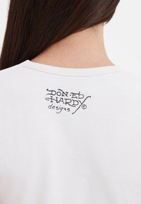 Hvid bomulds T-shirt med et sort, stiliseret logo trykt på den øverste del af ryggen. Blød tekstur og klassisk rund halsudskæring.