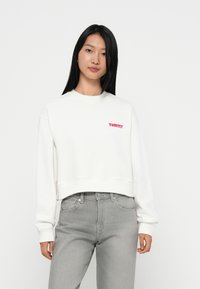Witte cropped sweatshirt met rood "Tommy" logo, lange mouwen en geribbelde manchetten, gecombineerd met lichtgrijze jeans. Soepele textuur, casual ontwerp.
