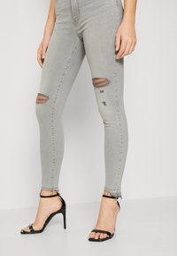 Mujer vestida con jeans skinny rasgados de color gris claro y sandalias de tacón negro, de pie contra un fondo liso.