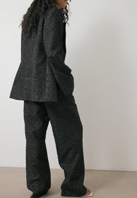 Zwarte textuur blazer met een fitted jasje en wijde broek met een subtiel reliëfpatroon, geplaatst tegen een neutrale achtergrond.