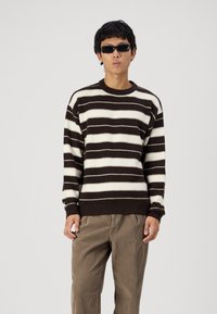 Brown and cream striped sweater with a round neckline and ribbed cuffs, paired with beige corduroy pants. Knitted texture, relaxed fit.