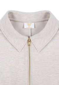 Ljus beige zip-up-overdel med utbrett krage, som har en guldfärgad dragkedja och mjukt, texturerat material. Etiketten visar varumärke och storleksinformation.