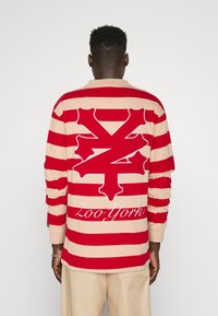 Long-sleeve T-shirt featuring red and beige horizontal stripes, large white logo on the back, and ribbed cuffs. Cotton fabric.