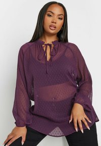 Blusa lilla trasparente con pois testurizzati, scollatura arricciata, dettaglio con fiocco e maniche lunghe a sbuffo. Abbinata a pantaloni neri.