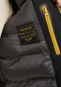 Interno di un giubbotto pieno grigio con un'etichetta nera caratterizzata da testo giallo, dettagli in zip dorati e una texture trapuntata.