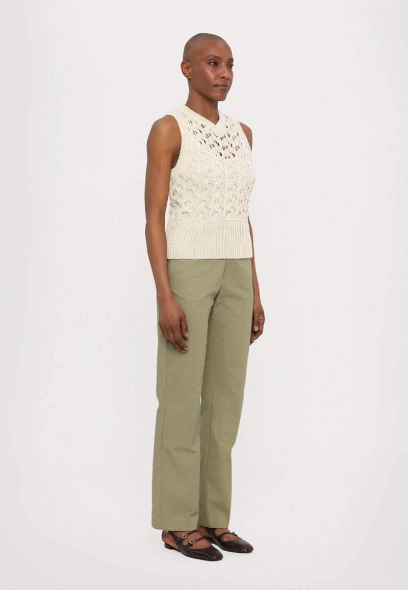 Débardeur en tricot sans manches de couleur crème avec un motif texturé, associé à un pantalon large vert sauge. Chaussures plates noires avec des accents en métal.
