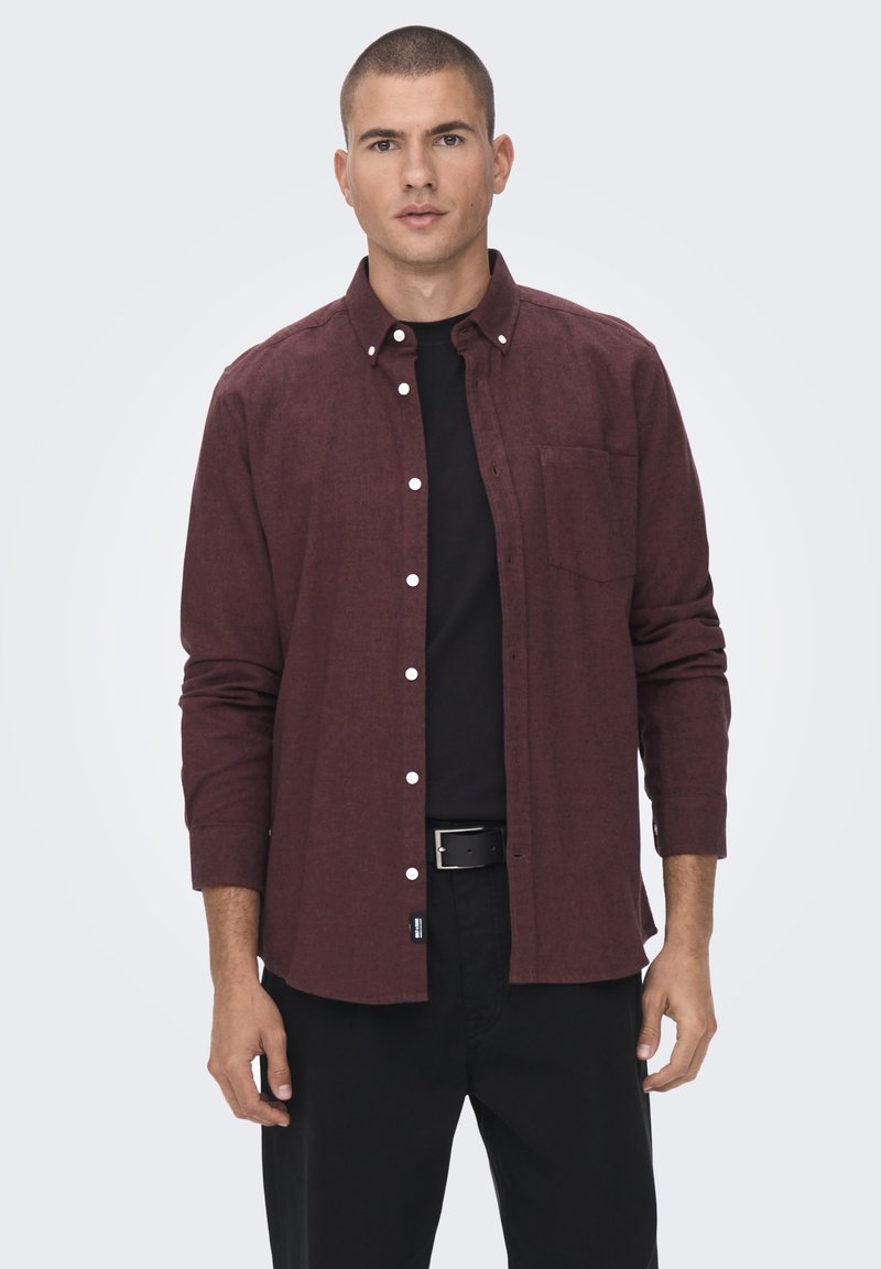 Chemise boutonnée bordeaux à manches longues, avec une poche frontale et des boutons blancs, portée par-dessus une chemise noire, assortie à un pantalon noir. Texture lisse.