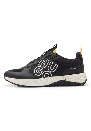 Baskets noires et blanches avec de grandes lettres blanches "HUGO" sur le côté, accents jaunes et semelle blanche mouchetée, vues du côté extérieur.