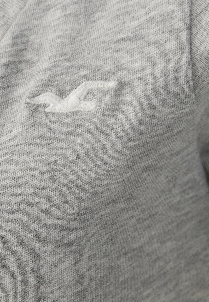 Jasnoszara bluza z teksturowanej tkaniny, z małym, białym haftowanym logo przypominającym ptaka po lewej stronie.