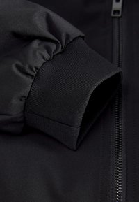 Close-up of a black jacket sleeve with elastic cuff and visible black zipper on smooth fabric.