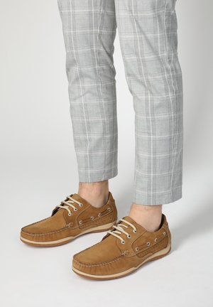 Lysebrune suede bådsko med en mokkasinlignende design, beige snørebånd og perforerede detaljer. Iført med grå ternede bukser.