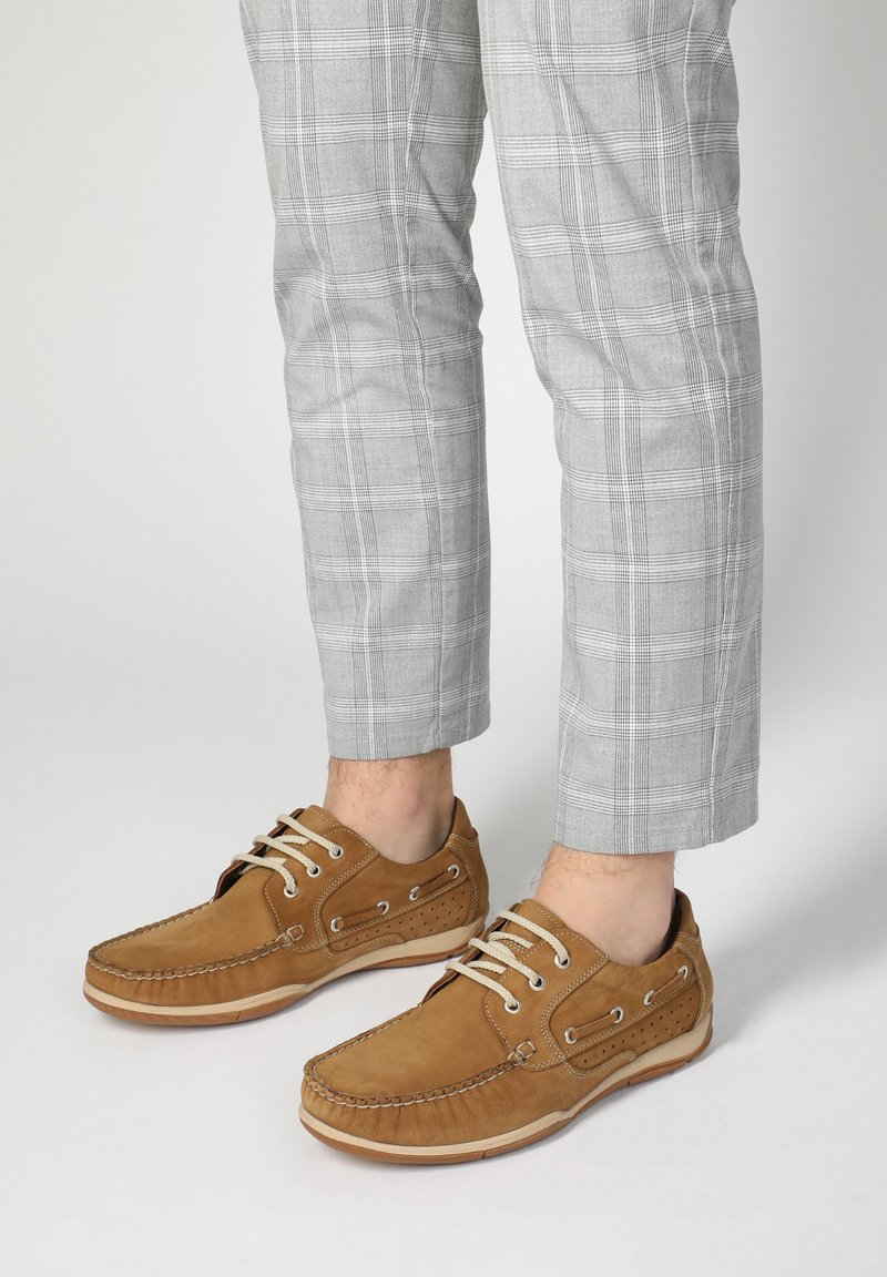Hellbraune Wildlederschuhe im Boots-Design mit Mokassin-Details, beige Schnürsenkel und durchbrochenen Akzenten. Getragen mit karrierten grauen Hosen.